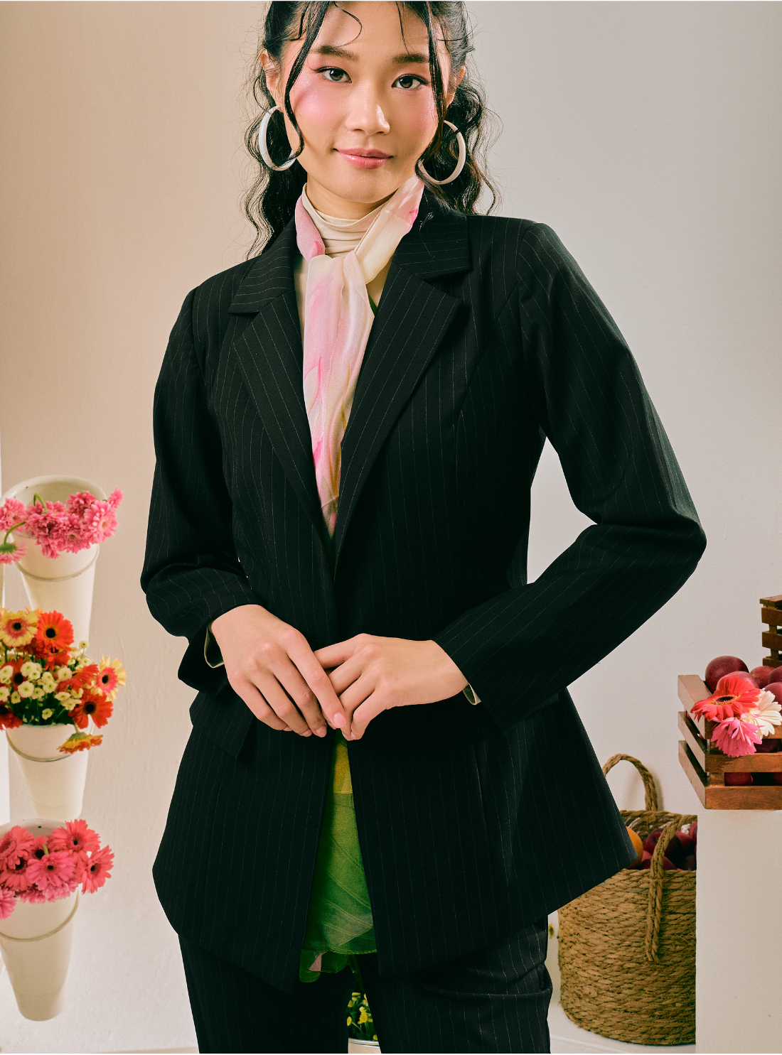 A woman wearing a black pinstriped blazer over a light pink scarf and green top, standing in a studio setting with decorative flowers and a basket.