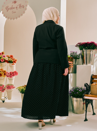 A woman viewed from behind wearing a black long-sleeve top and a black midi skirt with small white polka dots, standing in a styled indoor boutique setting with flowers and a bench.