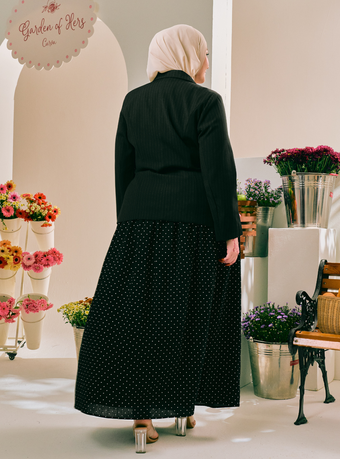 A woman viewed from behind wearing a black long-sleeve top and a black midi skirt with small white polka dots, standing in a styled indoor boutique setting with flowers and a bench.