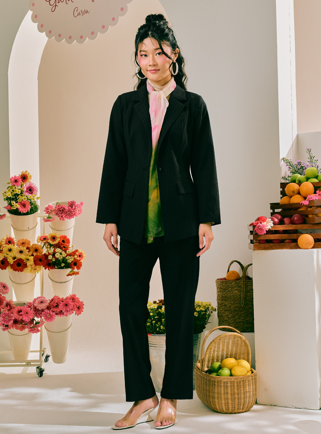 A woman stands in a styled indoor set wearing a black blazer and matching black pants over a pastel tie-dye shirt. The scene includes flower arrangements, fruit baskets, and a white arched backdrop.