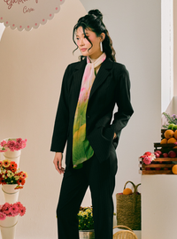A woman models a black two-piece suit with a vibrant tie-dye scarf in shades of green, yellow, pink, and white. She is standing in a styled indoor setting with decorative flowers and fruit baskets.