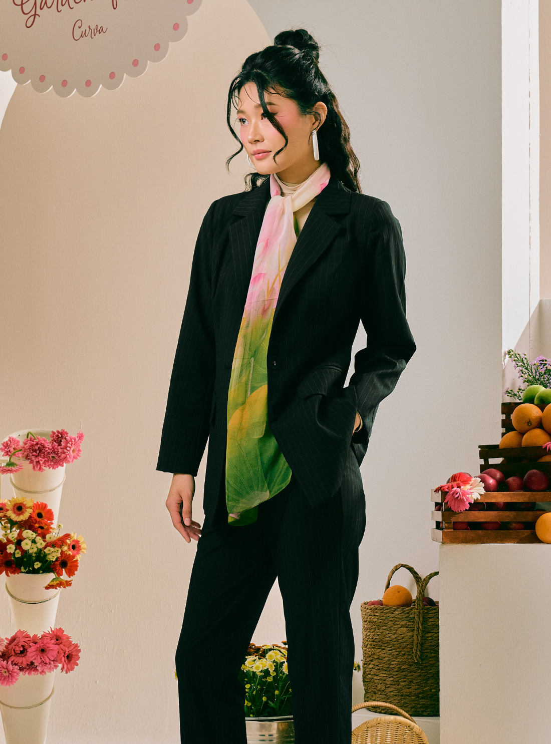 A woman models a black two-piece suit with a vibrant tie-dye scarf in shades of green, yellow, pink, and white. She is standing in a styled indoor setting with decorative flowers and fruit baskets.