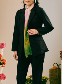 A model wearing a black pinstripe blazer and matching trousers, styled with a vibrant pink, green, and yellow tie-dye scarf, against a warm beige background with decorative flowers and a basket.