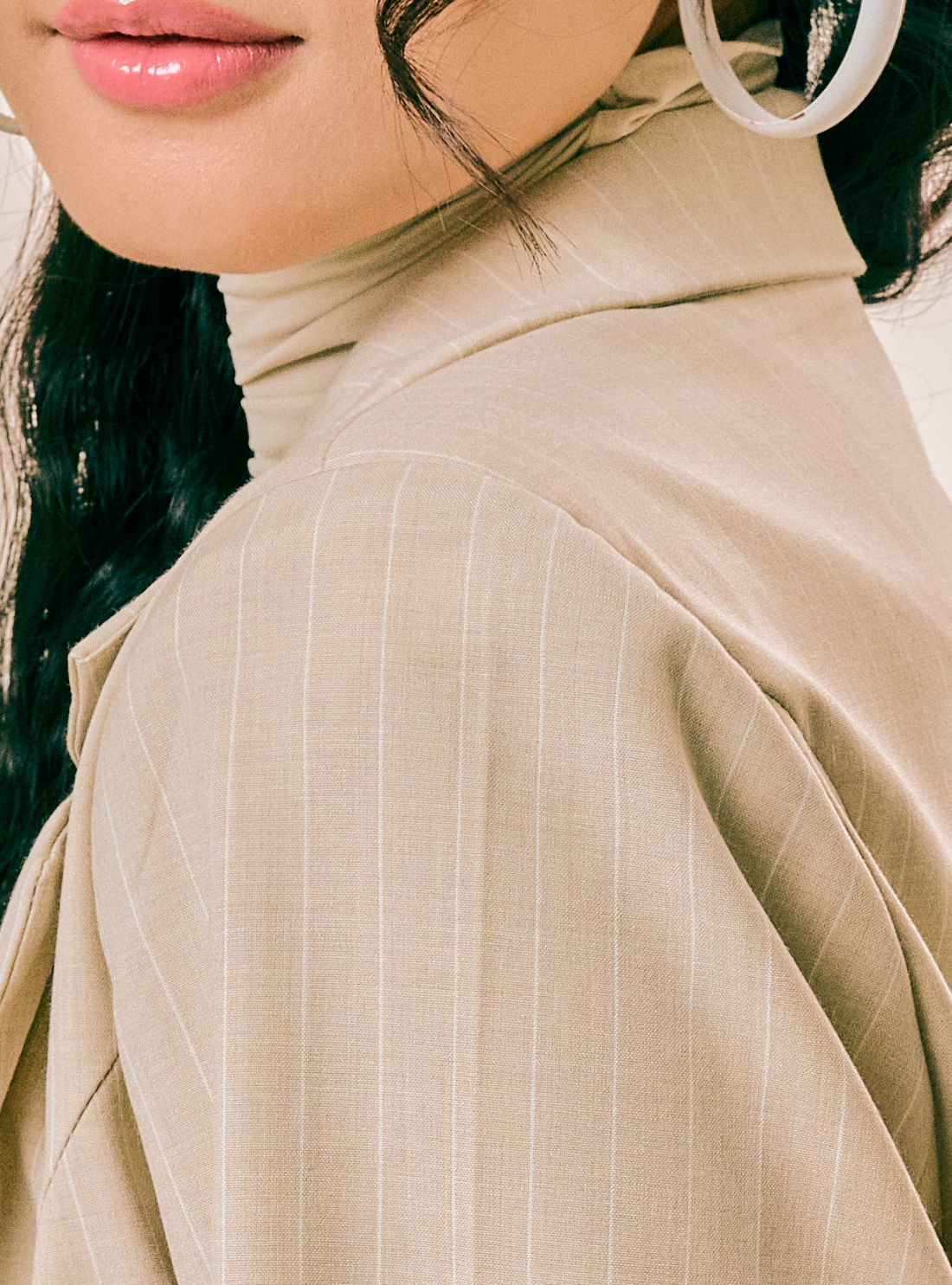 Close-up of a person wearing a beige or light tan high-neck top with subtle vertical pinstripes. The crop includes part of the face, hair, and a large hoop earring against a plain light background.