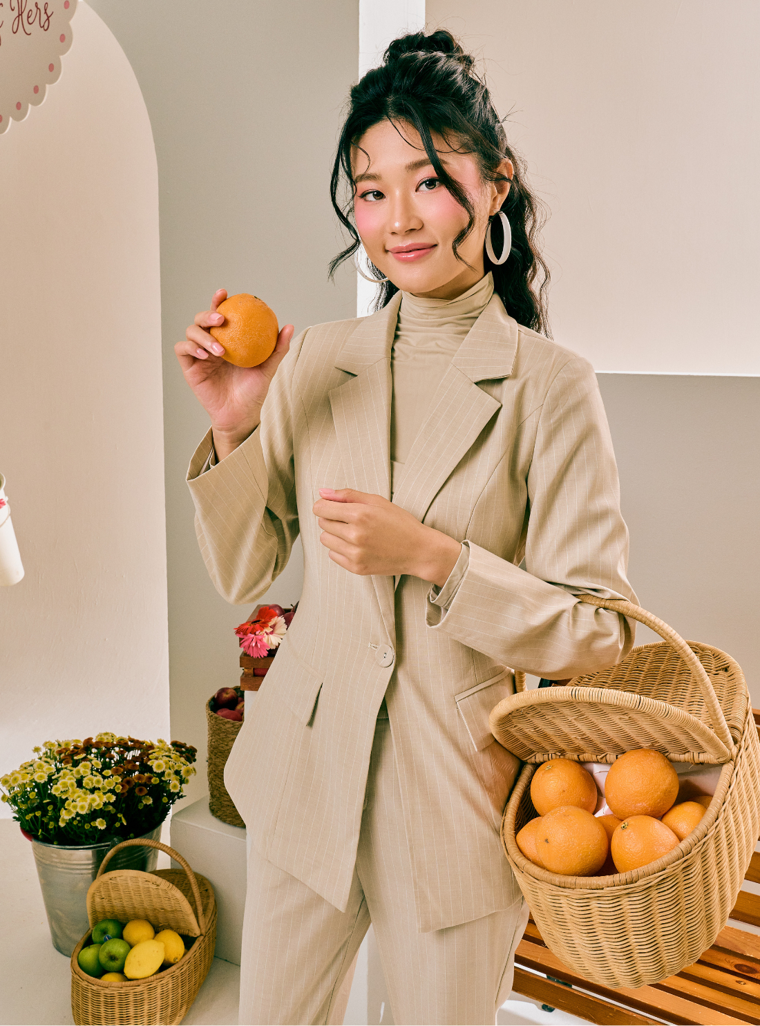 A woman wearing a beige two-piece blazer and turtleneck suit holds an orange in one hand and a woven basket of oranges in the other, standing in a brightly lit indoor setting with decorative baskets and flowers.