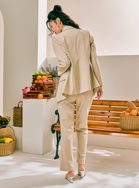 A woman wearing a beige two-piece suit with a structured blazer and straight-leg trousers, photographed from behind in a bright, modern indoor setting with a wooden bench and fruit baskets.
