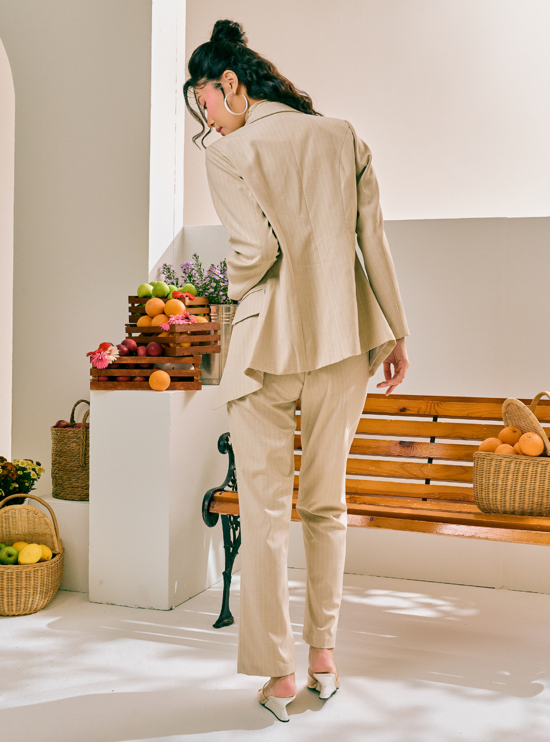 A woman wearing a beige two-piece suit with a structured blazer and straight-leg trousers, photographed from behind in a bright, modern indoor setting with a wooden bench and fruit baskets.