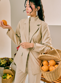 A woman wearing a beige double-breasted blazer and matching trousers over a beige mock neck top, holding an orange in a styled indoor setting with woven baskets of oranges.