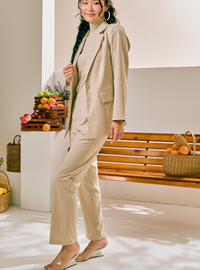 A woman models a beige two-piece suit consisting of a turtleneck top and matching cropped trousers, styled in a bright, modern indoor setting with a wooden bench and fruit baskets.