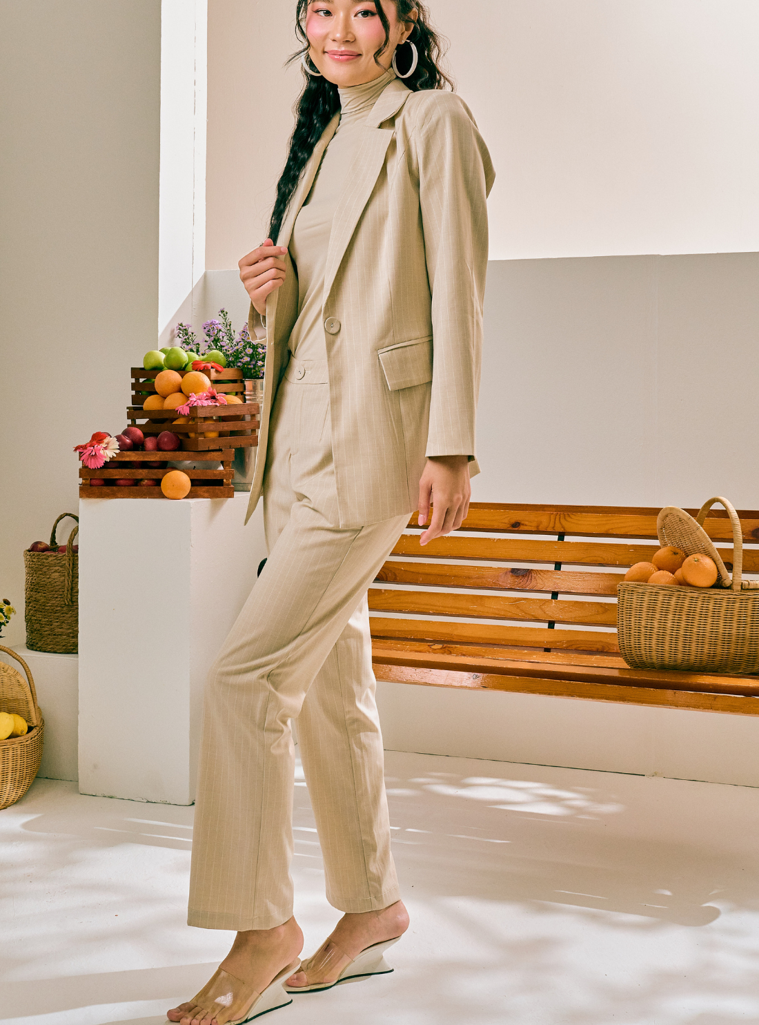 A woman models a beige two-piece suit consisting of a turtleneck top and matching cropped trousers, styled in a bright, modern indoor setting with a wooden bench and fruit baskets.