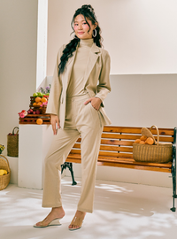 A woman models a beige two-piece suit consisting of a long-sleeve blazer and matching high-neck top and trousers, paired with beige heeled sandals in a bright, modern indoor setting with a wooden bench and fruit baskets.