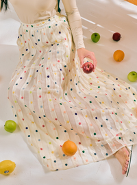 A woman seated on a white floor wearing a long cream skirt with multicolored polka dots and a matching cream long-sleeve top. Fruit is scattered around her on the floor.