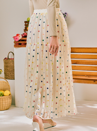 A model wearing a cream-colored, tiered tulle maxi skirt with colorful polka dots, paired with a beige long-sleeve top and white slide sandals, standing in a bright indoor setting.