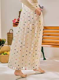 A woman wearing a long cream-colored sheer maxi skirt with colorful embroidered polka dots, standing in a bright indoor setting with a wooden bench and baskets in the background.