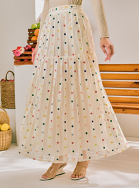 A model wearing a cream-colored, tiered tulle maxi skirt with a colorful polka dot pattern, paired with beige flat sandals in a bright indoor setting.