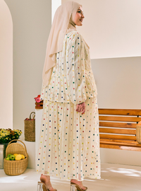 A woman wearing a cream-colored two-piece modest outfit with a small multicolor polka dot pattern, paired with a matching beige hijab, standing in a bright, modern interior setting.