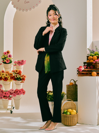 A woman models a black two-piece blazer and trouser set with a pink shirt and a green-yellow patterned scarf, standing in a bright, styled studio with flowers and fruit baskets.