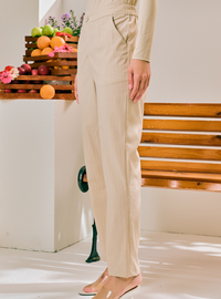 A person wearing light beige wide-leg trousers with a matching long-sleeve top, standing barefoot in a bright indoor setting with a wooden bench and fruit crates in the background.
