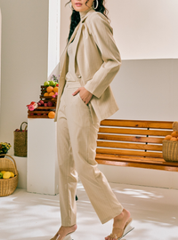 A woman models a beige two-piece suit consisting of a long-sleeve blazer and matching wide-leg trousers, styled with clear strap heels in a bright, modern indoor setting.