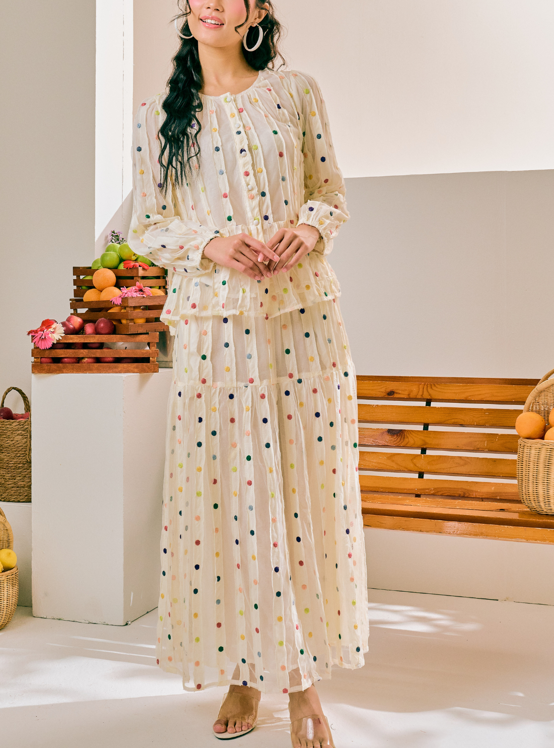 A woman models a long-sleeve, tiered maxi dress with a cream base and small multicolored polka dots. The dress features a gathered, ruffled neckline and a flowing skirt.