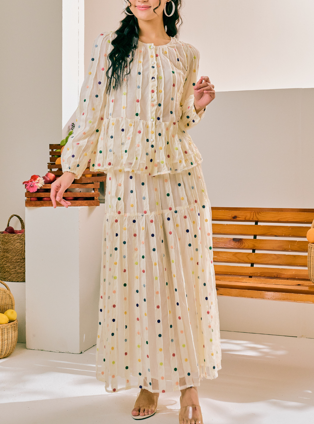 A woman models a two-piece matching set consisting of a long-sleeve ruffled blouse and a long pleated maxi skirt, both in a cream-colored sheer mesh fabric with a colorful polka dot pattern. The setting is a bright, modern indoor space with a wooden bench and white columns.