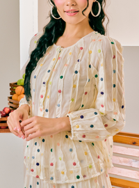 A woman wearing a sheer cream-colored long-sleeve blouse with colorful embroidered polka dots, paired with large gold hoop earrings, in a brightly lit indoor setting.