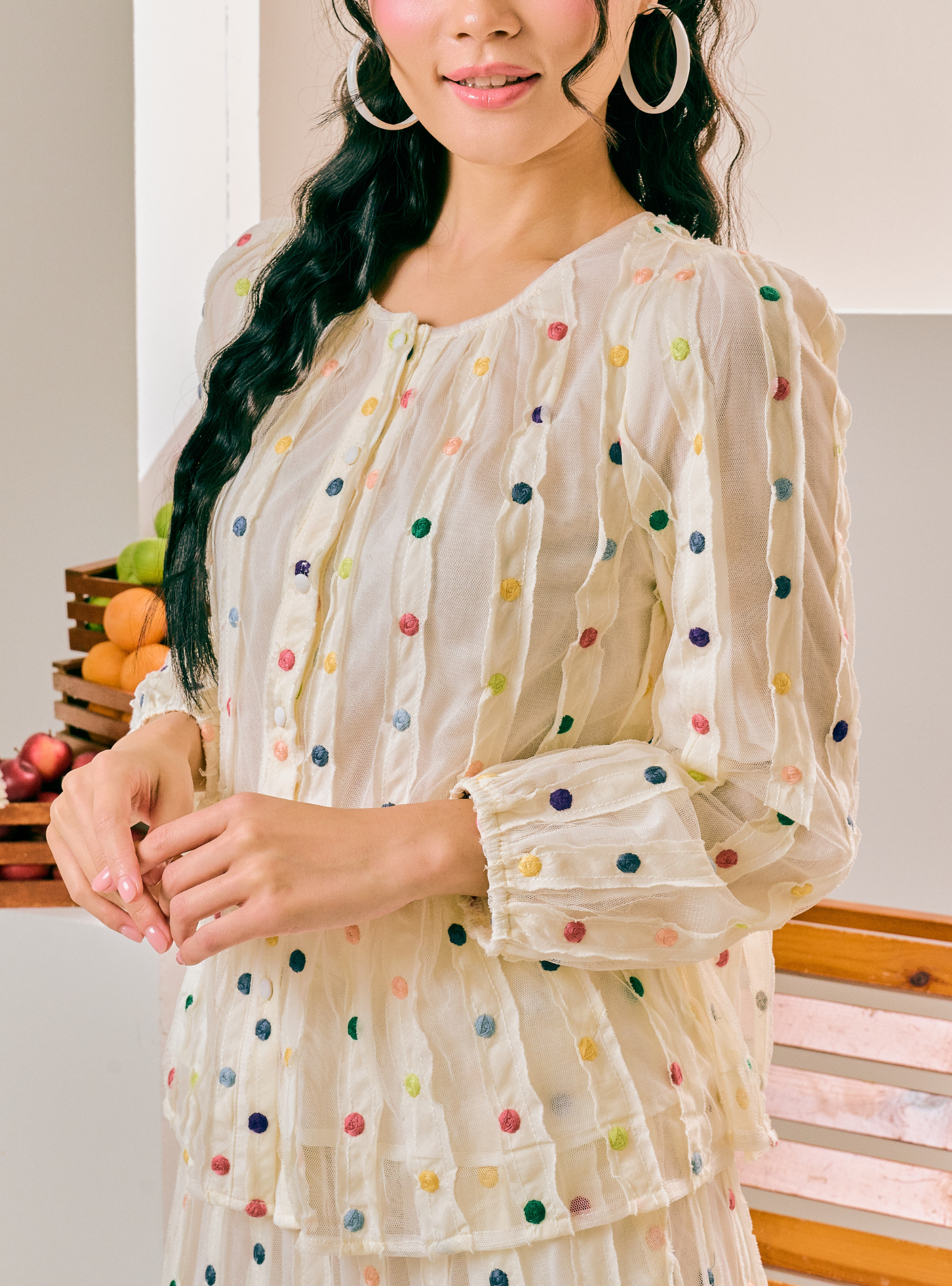 A woman wearing a sheer cream-colored long-sleeve blouse with colorful embroidered polka dots, paired with large gold hoop earrings, in a brightly lit indoor setting.