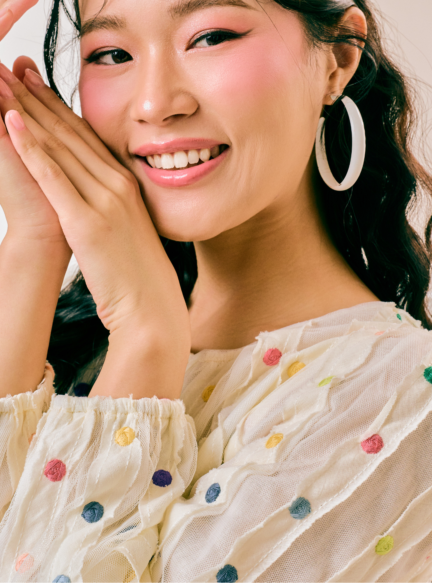 A close-up lifestyle shot of a smiling woman with dark hair wearing a cream-colored, ruffled blouse with colorful embroidered polka dots and a large white hoop earring.
