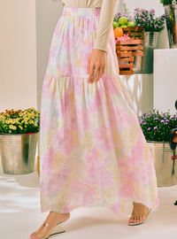 A woman wearing a long tiered maxi skirt with a pastel pink, yellow, and white tie-dye pattern and eyelet embroidery, paired with a beige long-sleeve top and clear wedge sandals.