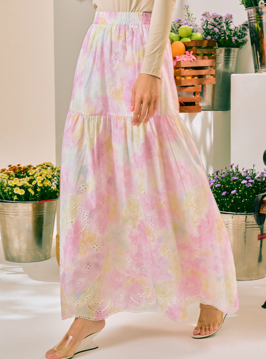 A woman wearing a long tiered maxi skirt with a pastel pink, yellow, and white tie-dye pattern and eyelet embroidery, paired with a beige long-sleeve top and clear wedge sandals.