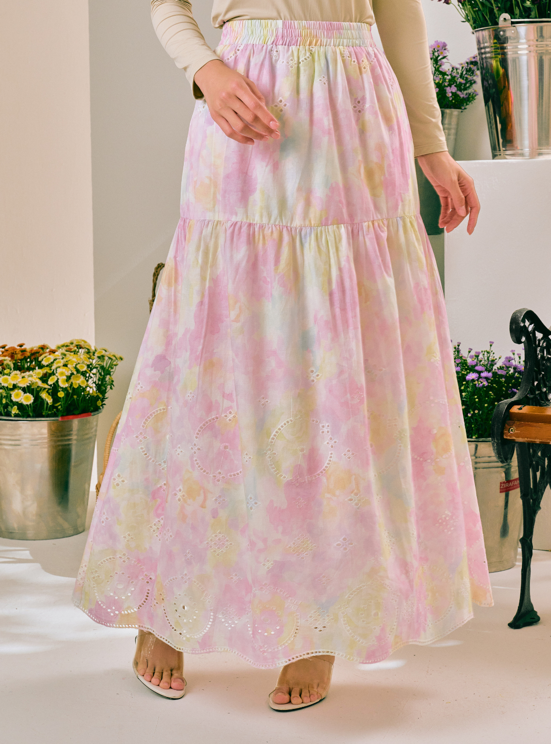 A woman wearing a long tiered maxi skirt with a pastel pink, yellow, and white tie-dye pattern, paired with beige heeled sandals in an indoor setting.