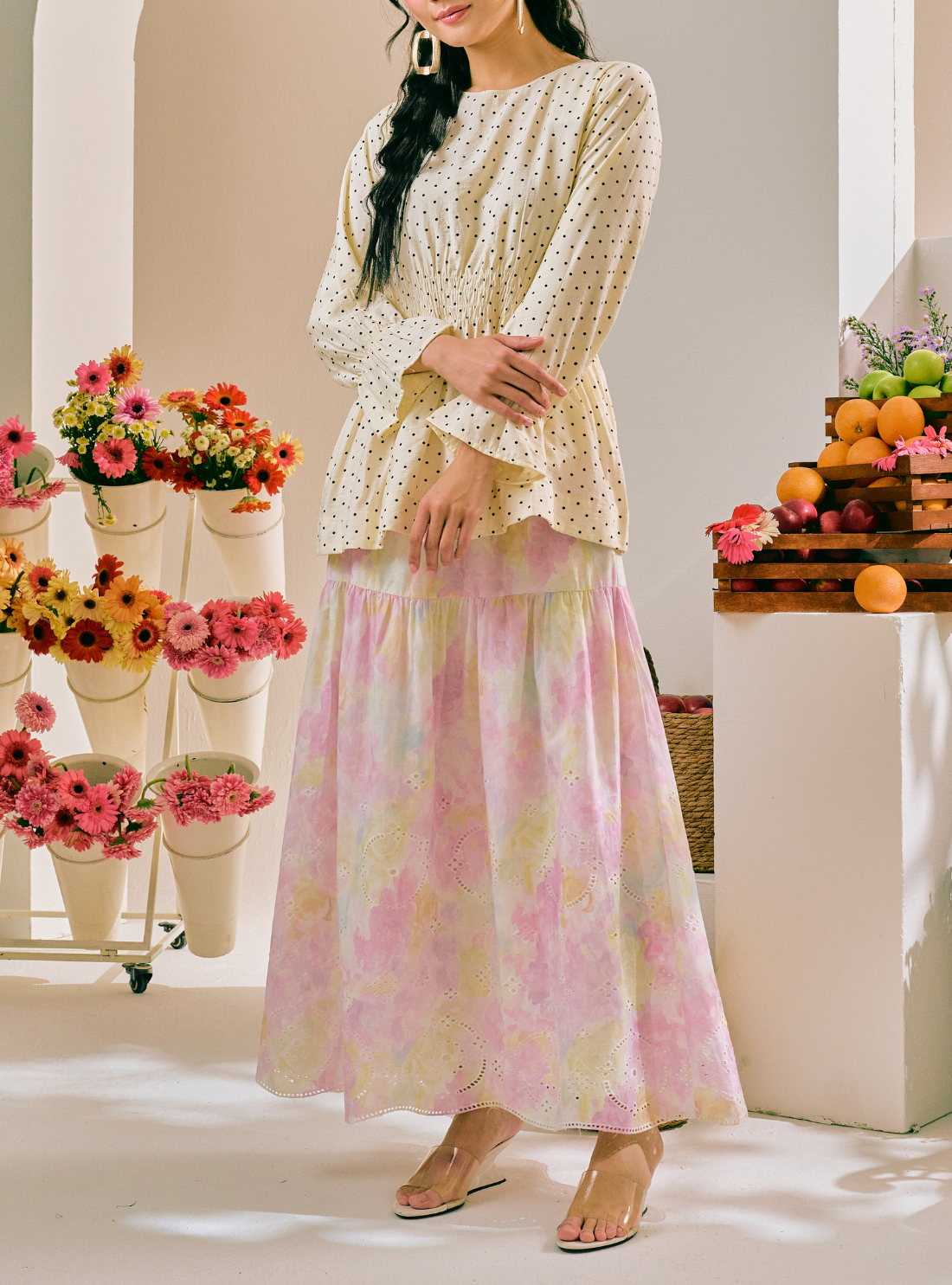 A woman models a two-piece outfit consisting of a cream-colored long-sleeve blouse with small black polka dots and a matching tiered maxi skirt with a pink, yellow, and white floral pattern. She is standing in a brightly lit indoor setting decorated with flowers and fruit.