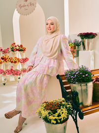 A woman wearing a pastel pink and yellow tie-dye two-piece modest outfit with a matching hijab, posing on a wooden bench in a flower shop setting.