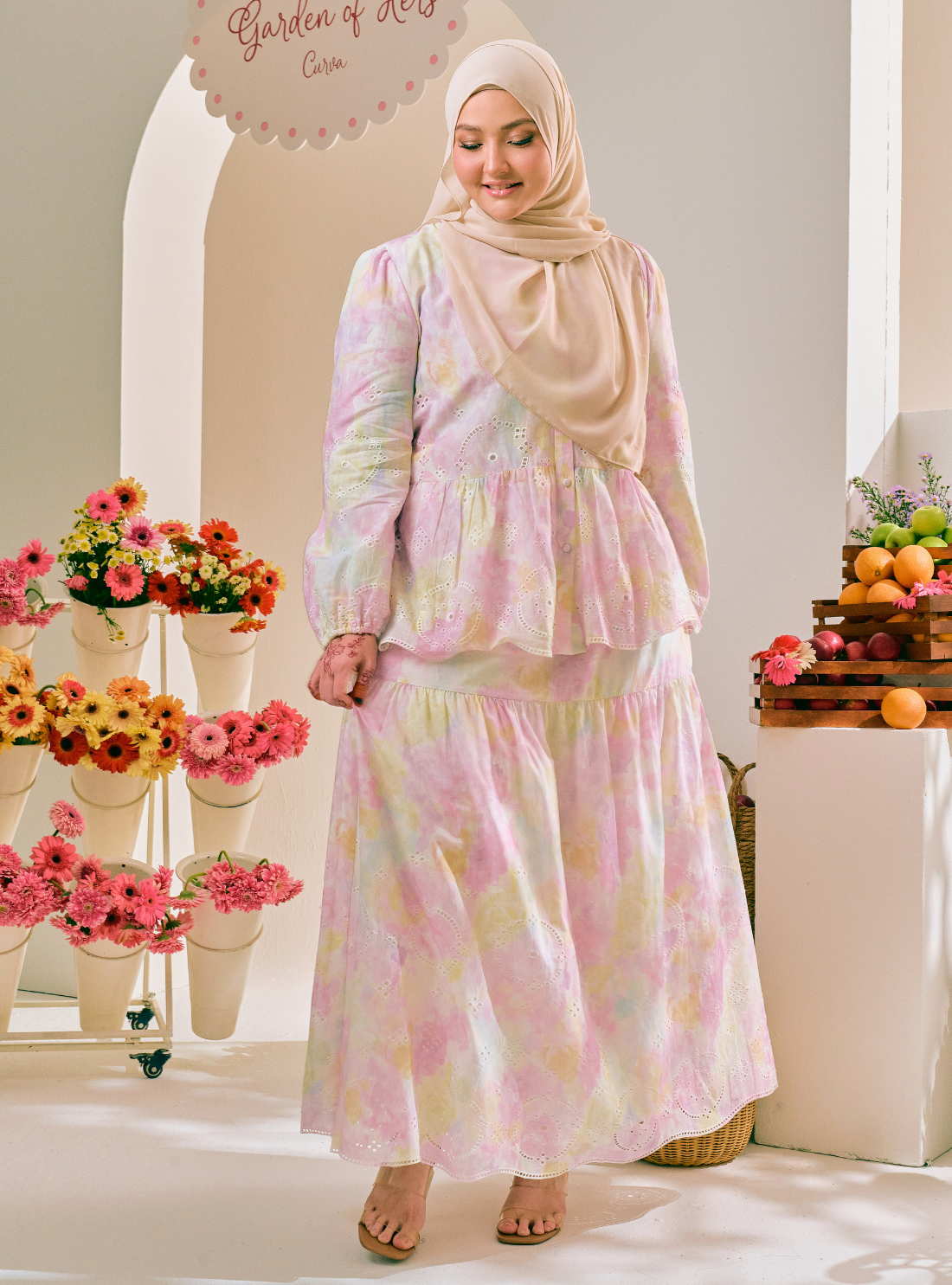A woman wearing a pastel multicolor floral print modest dress with a matching hijab, standing in a bright, decorated studio setting with flowers and fruit props.