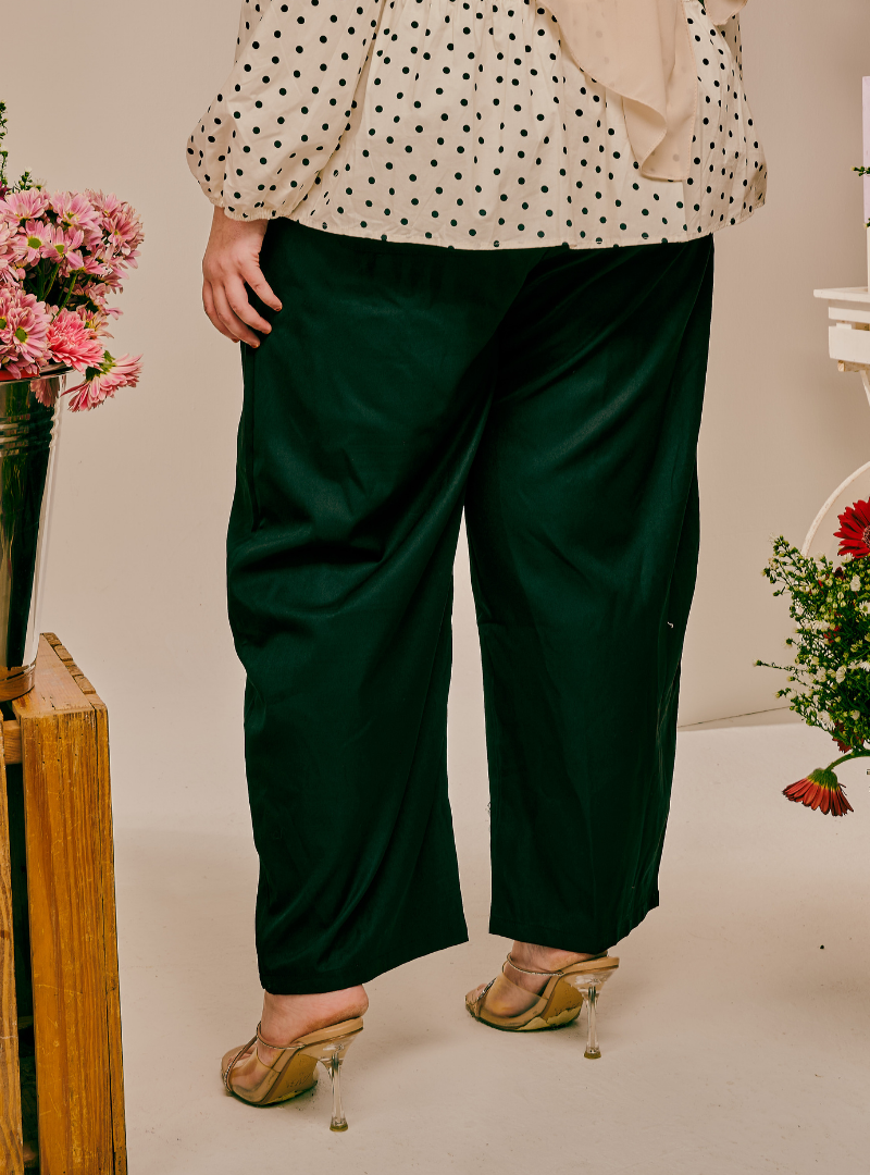 A person wearing dark green wide-leg cropped pants, a cream blouse with black polka dots, and clear-strap high heels, shown from the waist down in a styled indoor setting with flowers.