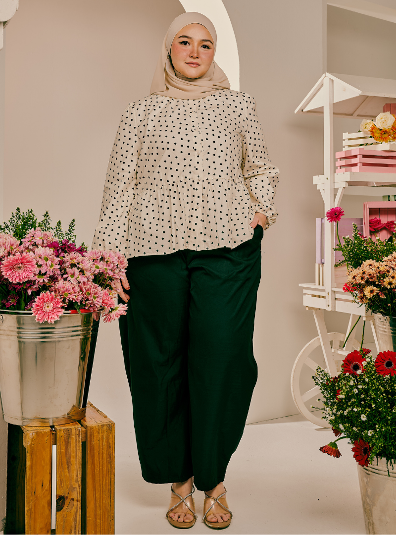 A woman modeling a long-sleeve cream blouse with black polka dots, paired with dark green wide-leg pants and a beige hijab, standing in a styled studio setting with flowers and a white decorative cart.
