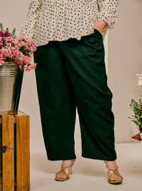 A person wearing dark forest green wide-leg cropped pants, paired with a cream blouse with black polka dots and metallic strappy heels, standing in a studio setting with decorative flowers.