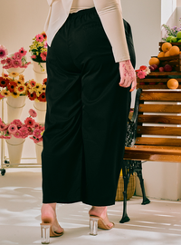 A person wearing black wide-leg cropped pants with an elastic waist, paired with a beige long-sleeve top and clear block-heel mules, standing in a brightly lit studio with flowers and a wooden bench in the background.