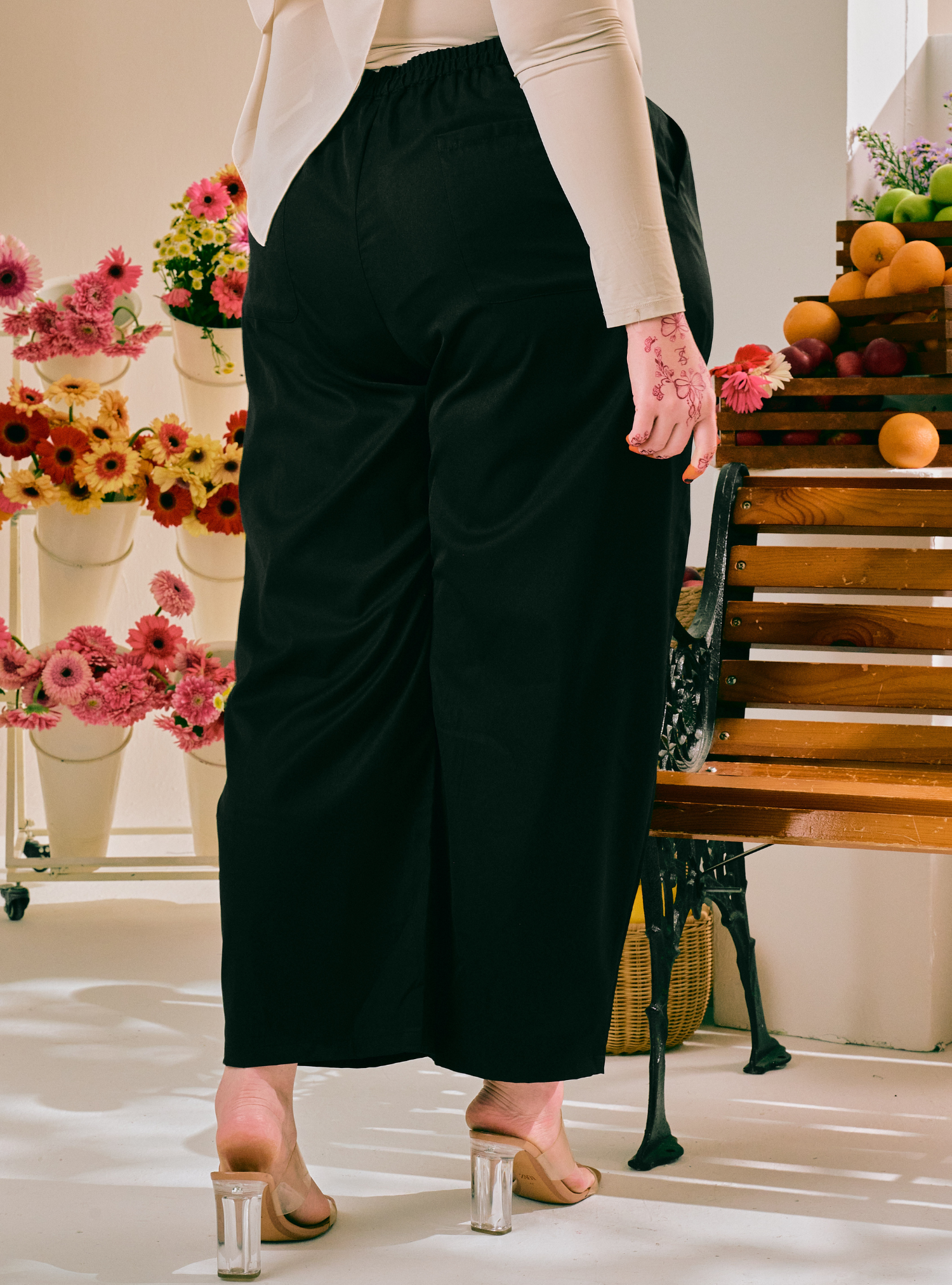 A person wearing black wide-leg cropped pants with an elastic waist, paired with a beige long-sleeve top and clear block-heel mules, standing in a brightly lit studio with flowers and a wooden bench in the background.