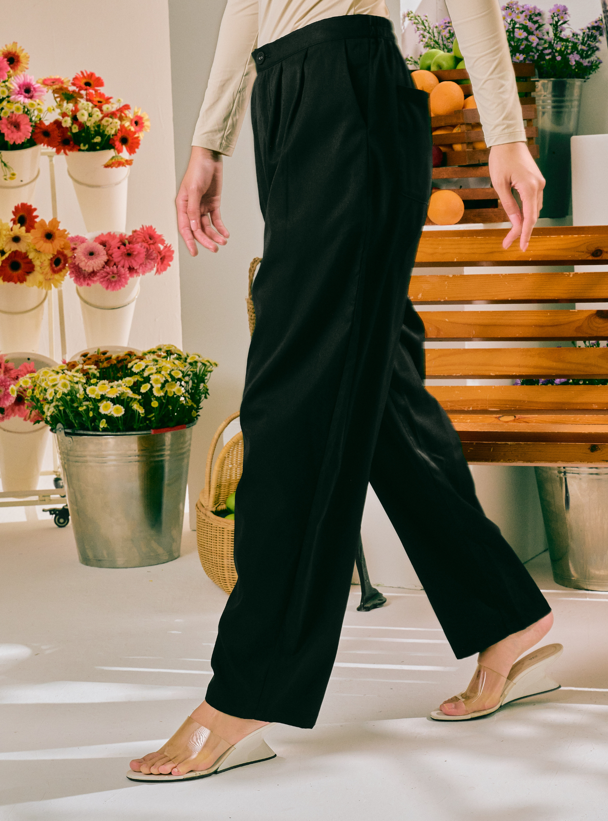 A person wearing black wide-leg cropped pants and clear wedge sandals, shown from the waist down in a bright indoor setting with flowers, fruit, and a wooden bench in the background.