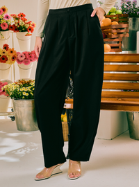 A person wearing black wide-leg cropped pants with a pleated front, paired with a light beige top and clear heeled sandals, standing in a bright indoor setting with flowers and wooden furniture in the background.