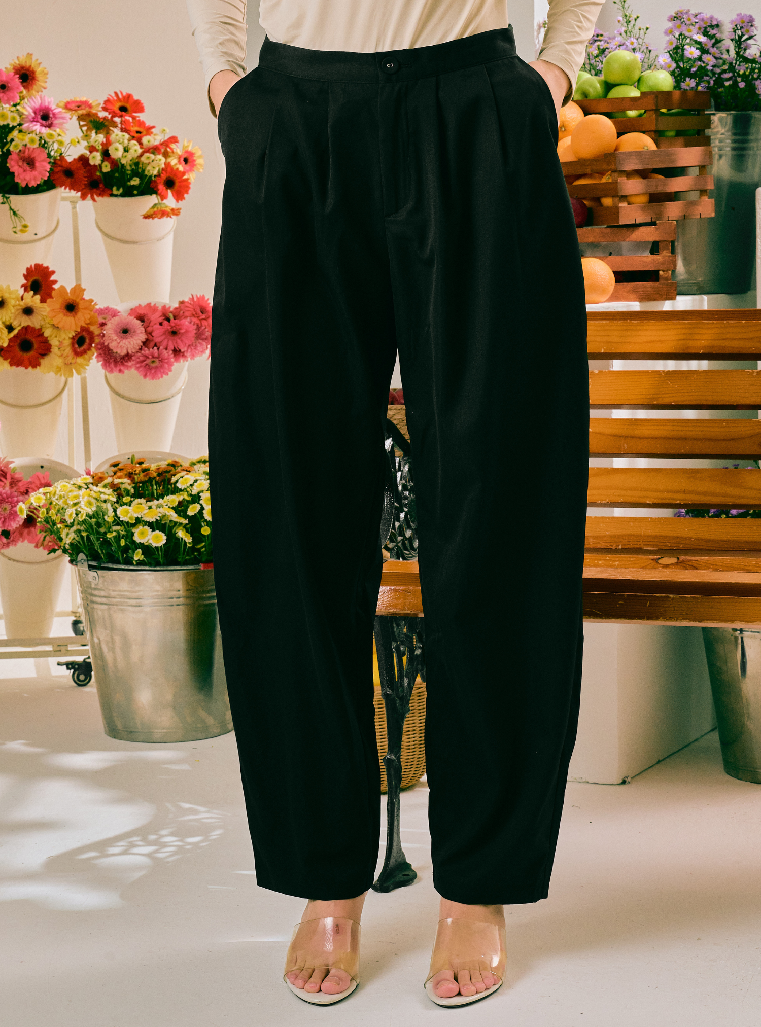 A person wearing black wide-leg pleated trousers with a cream top and clear high-heeled sandals, standing in a bright shop or studio setting with flowers and wooden furniture in the background.