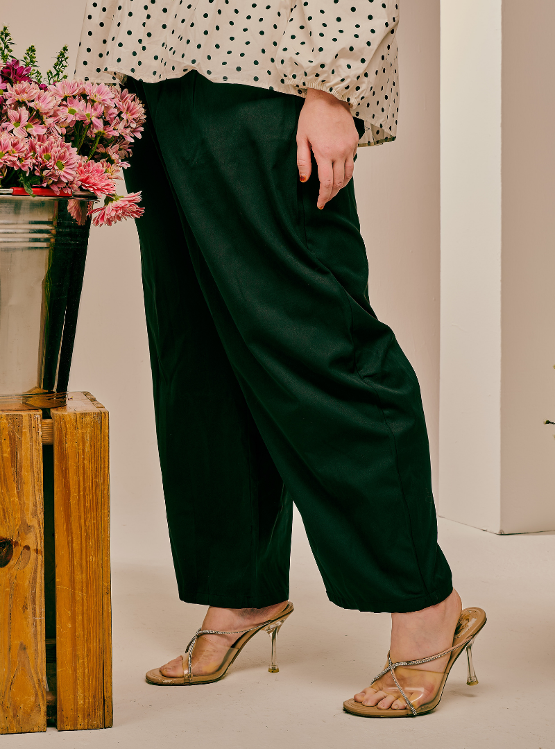 A person wearing dark forest green wide-leg cropped pants, paired with a cream blouse with black polka dots and clear high-heeled sandals, standing next to a wooden crate with pink flowers.