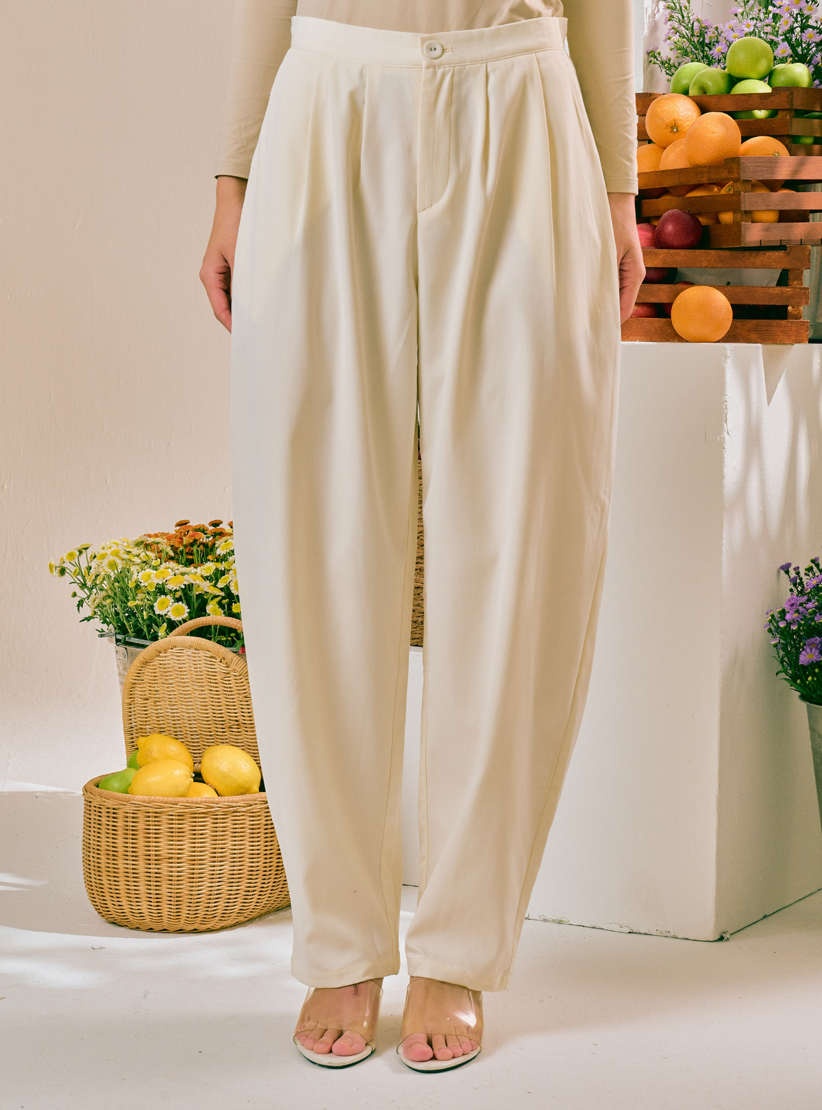 A person wearing high-waisted, wide-leg cream pants with a button closure, shown from the waist down in a styled indoor setting with baskets of fruit and flowers.