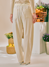 A person shown from the back wearing cream-colored wide-leg trousers with an elastic waistband and large patch pockets, standing in a styled studio setting with baskets of fruit and flowers.