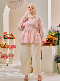 A woman wearing a light pink long-sleeve peplum top with a small white polka dot pattern, cream wide-leg pants, and a beige hijab, standing in a bright studio set decorated with flowers and fruit.
