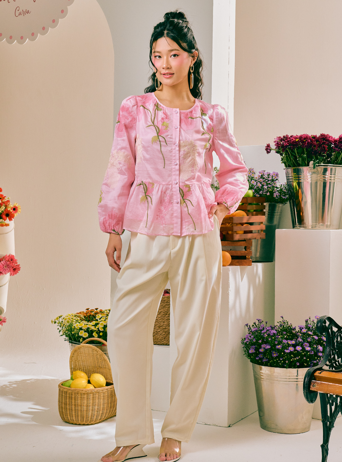 A woman models a pink long-sleeve button-up blouse with delicate floral embroidery and a peplum waist, paired with cream-colored wide-leg trousers, in a styled studio setting with potted plants and baskets.