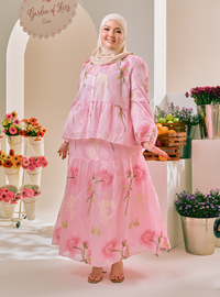 A woman wearing a light pink tiered maxi dress with a floral print and a matching hijab, standing in a bright, styled indoor setting with flowers and fruit.