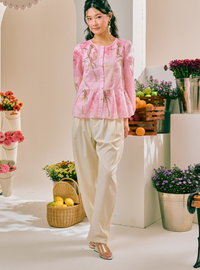 A woman models a light pink embroidered long-sleeve blouse with a peplum waist, paired with cream-colored trousers and metallic flat shoes, set against a bright, floral studio backdrop.