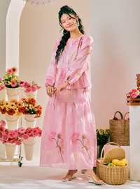 A woman models a light pink two-piece dress set with floral embroidery in a styled studio setting decorated with flowers and baskets.